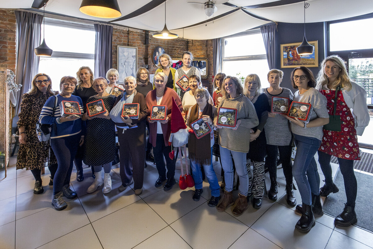 Około dwudziestu kobiet pozuje wspólnie w restauracji, trzymając puszki z piernikami przygotowanymi podczas spotkania „Oddech dla Mam”.